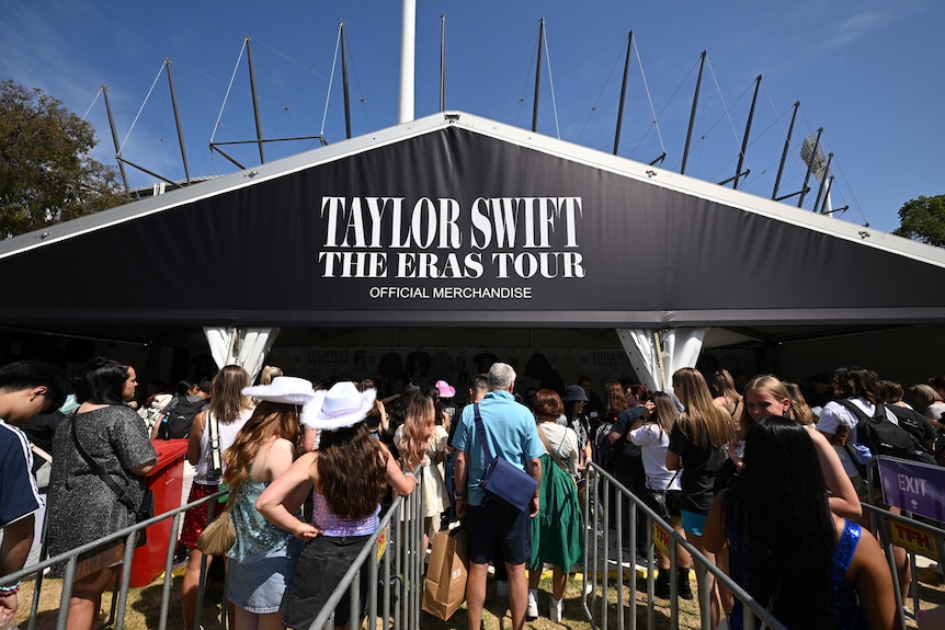 Fans in costume line in a queue to buy merchandise with a black 'Taylor Swift The Eras Tour' sign