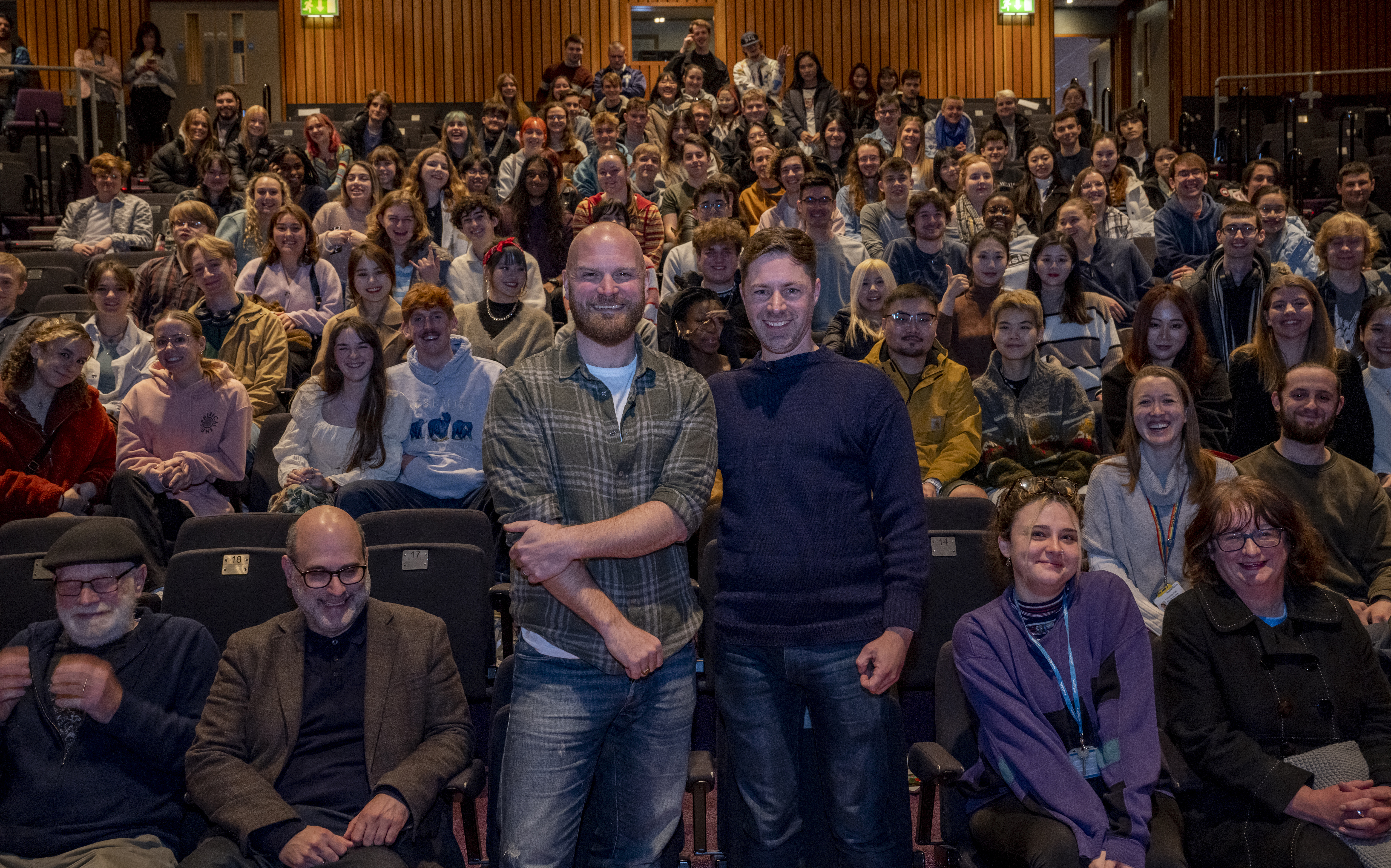 Will and Dan standing in front of students sat in the auditorium, smiling to camera