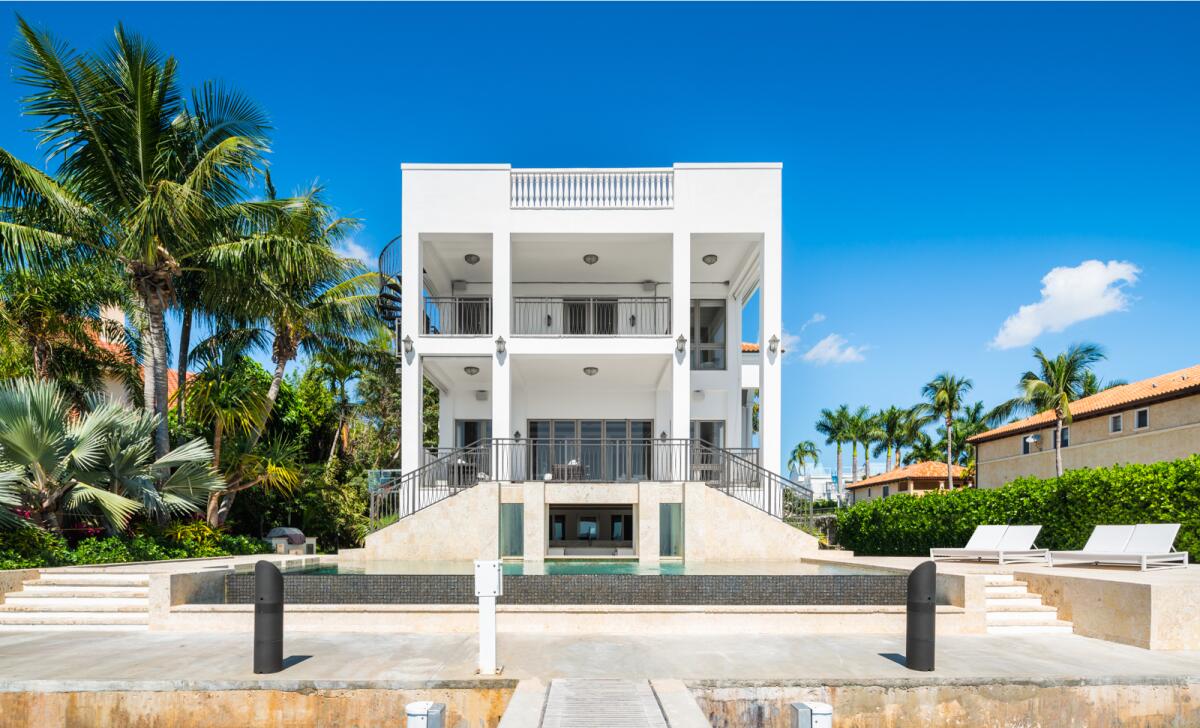 A view from the dock shows the rear of the mansion, an infinity pool and surrounding palm trees.